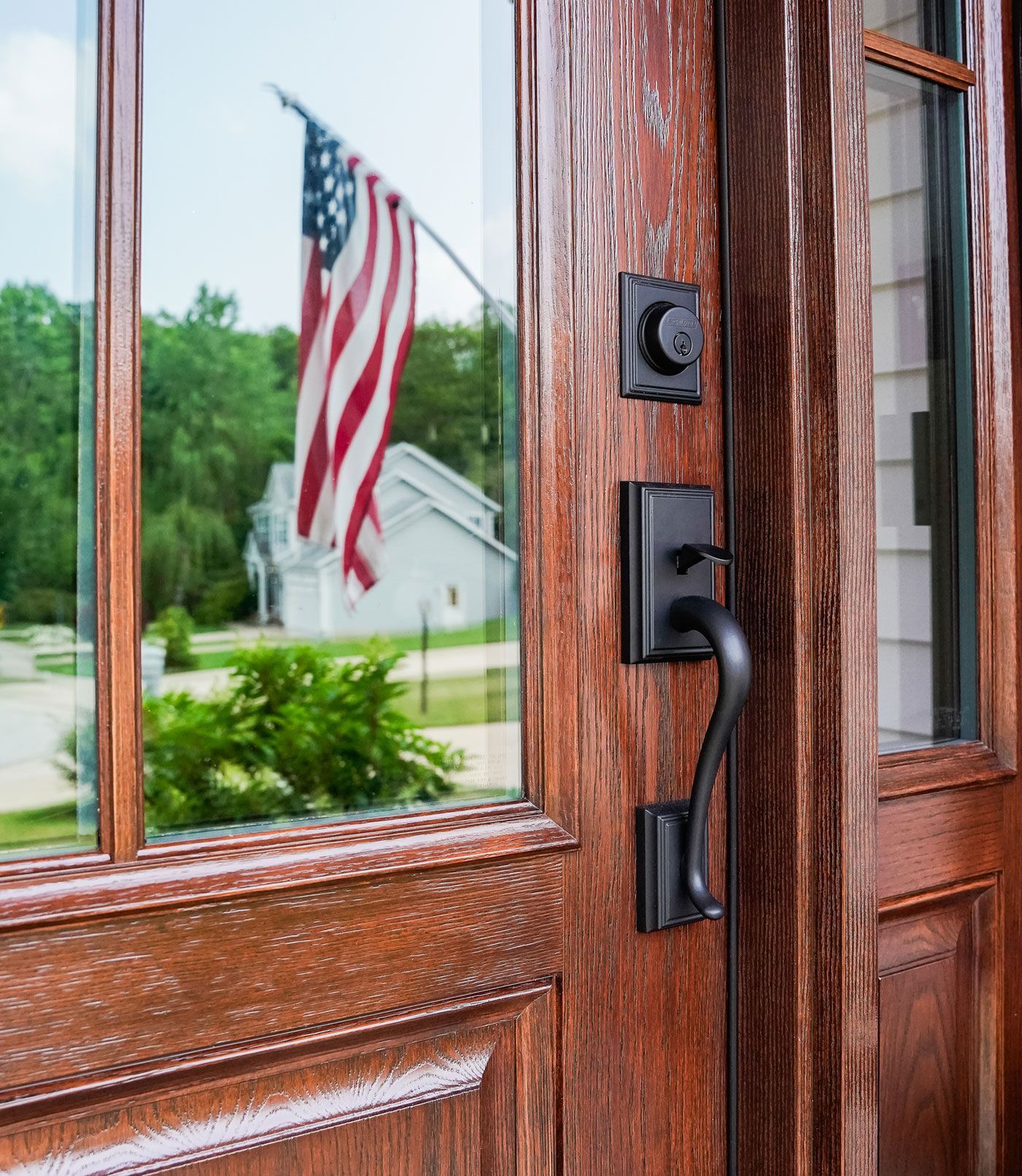 Close-up of a wooden front door with black handle, reflecting an American flag and house outside.