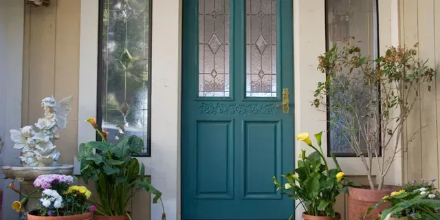 Teal front door with sidelights, flanked by potted flowers and greenery on a porch