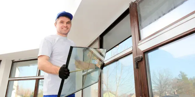 Worker in blue cap holding a window pane next to a house exterior