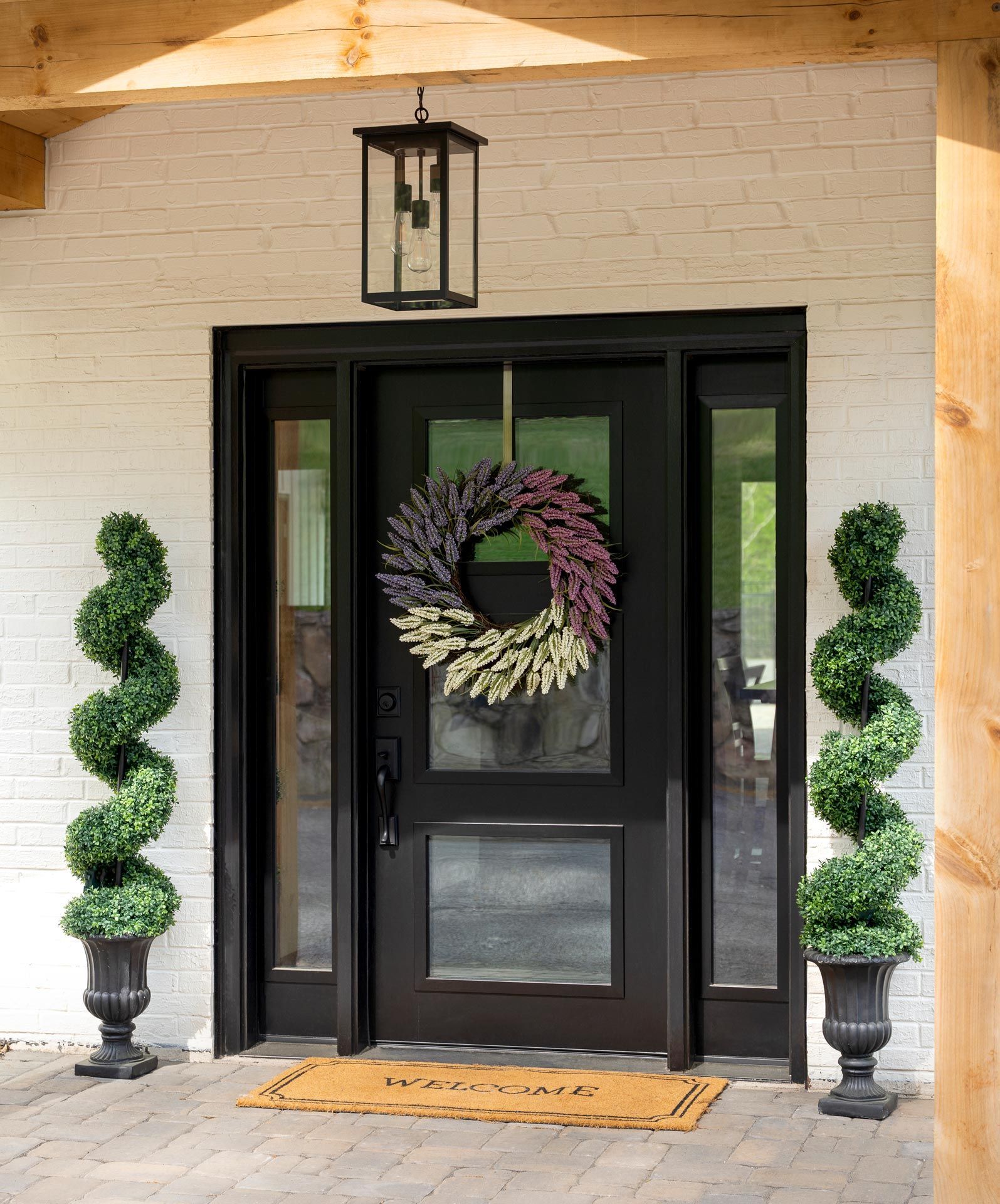 Front porch with black double doors, wreath, lantern, and two spiral topiary plants.