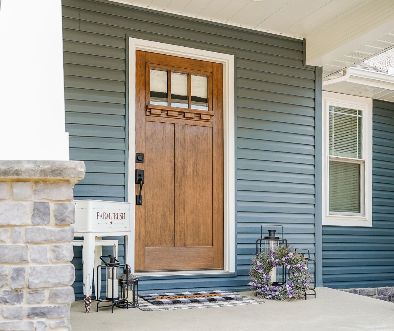 Front porch with a wood front door, blue siding, stone wall, lanterns, and a wreath beside a window.