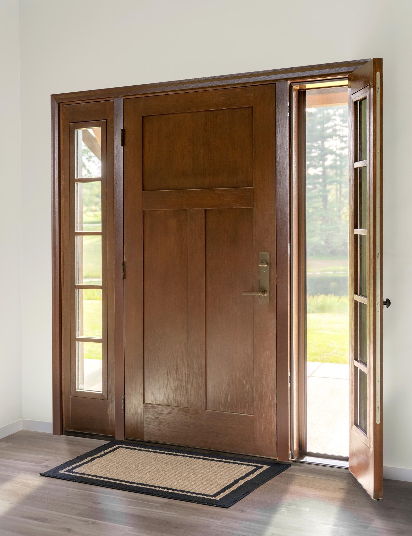 Wooden front door with glass sidelights and one open side door, viewed from indoors with a doormat.