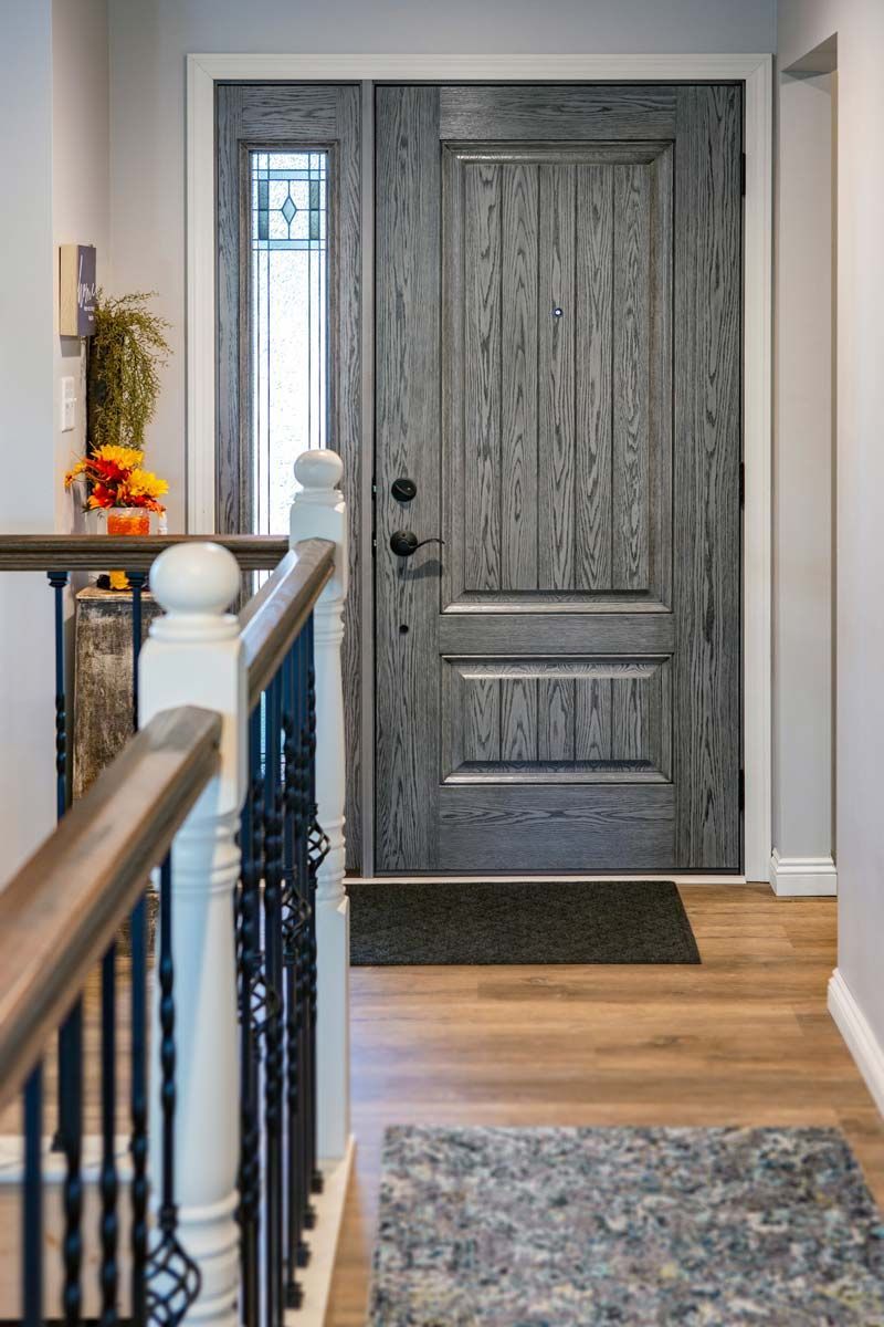 Gray front door in a hallway with wood floors, a stair railing, and a patterned rug.