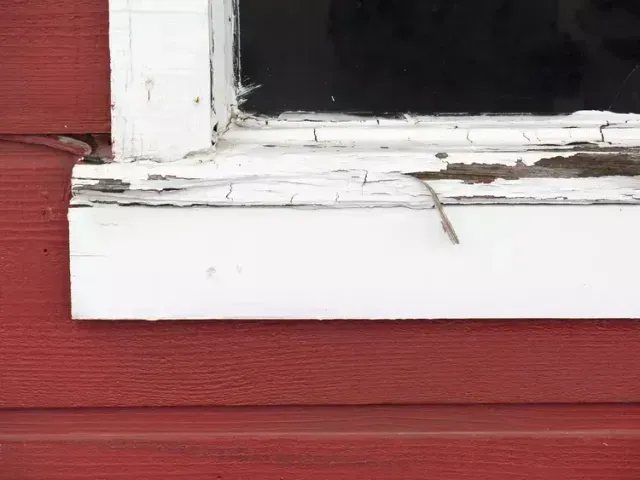 Close-up of a weathered white window frame on a red wall with peeling paint and a broken pane