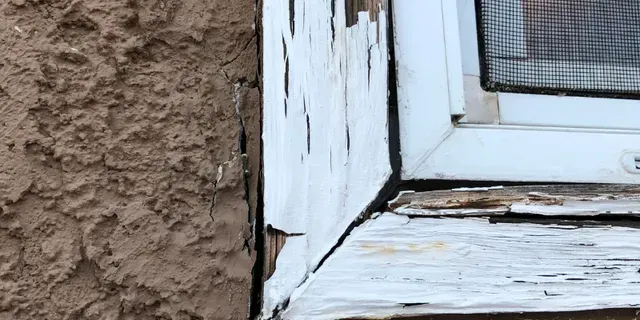 Close-up of peeling white window frame beside rough brown stucco wall and cracked sill