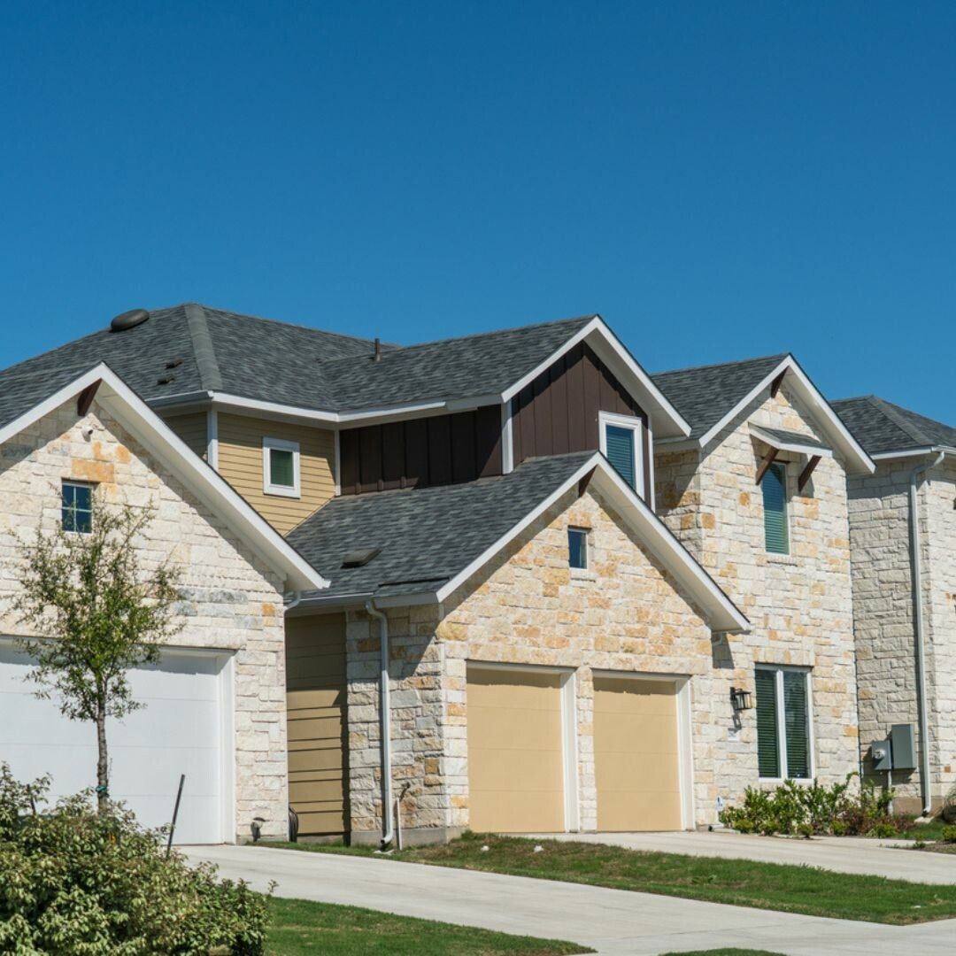 Two-story stone house with multiple gables and two garage doors, against a clear blue sky.