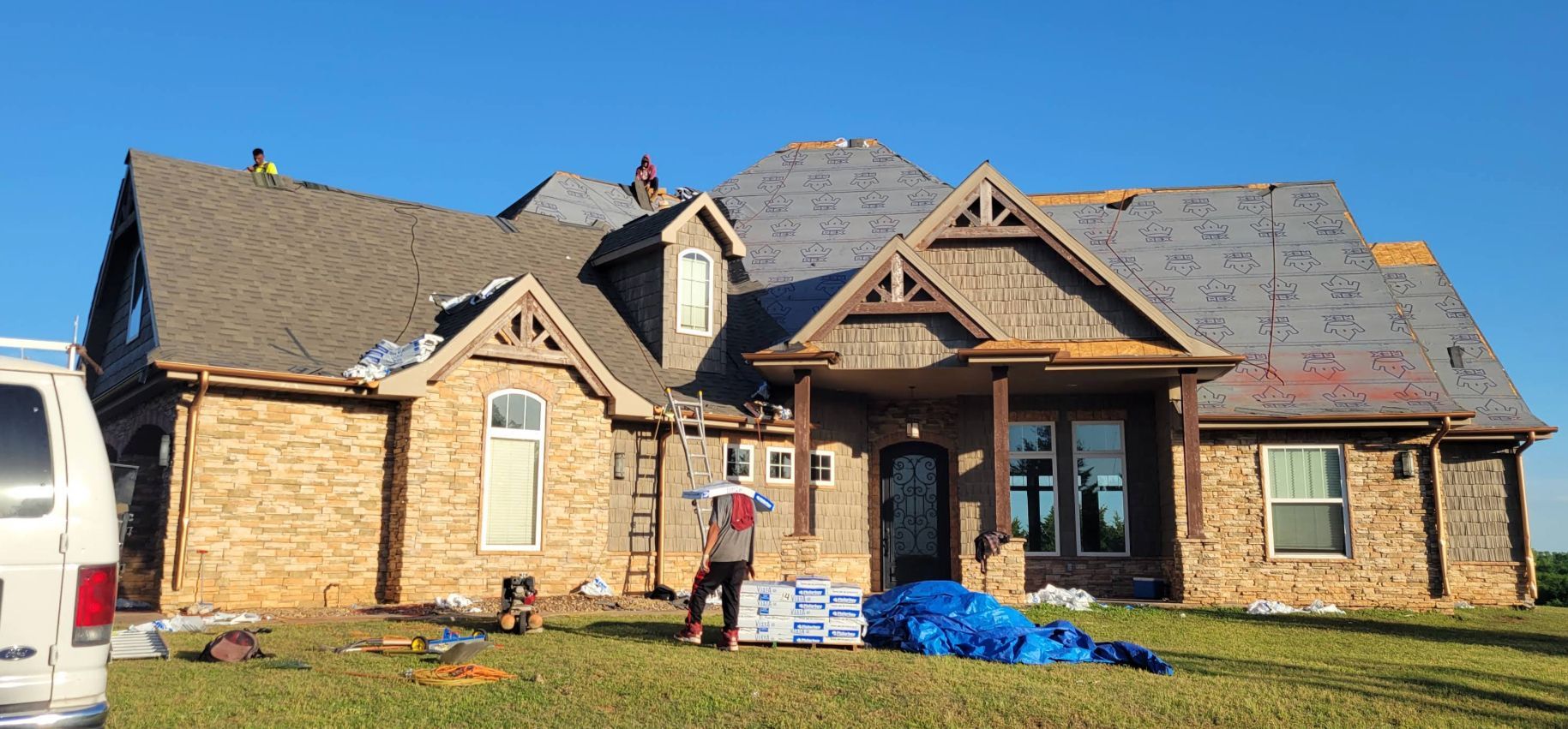 Roofers working on a house with brown brick and gray shingle roof. Blue sky and grass in front.
