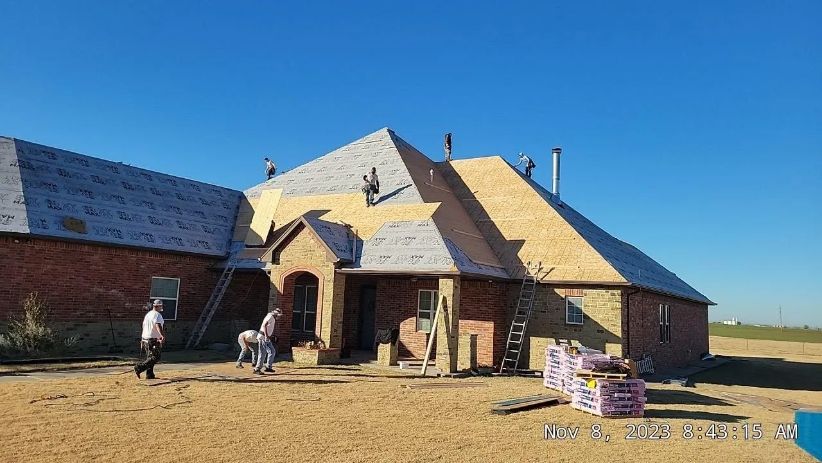 Roofers working on a brick house under a blue sky, with stacks of materials and ladders present.