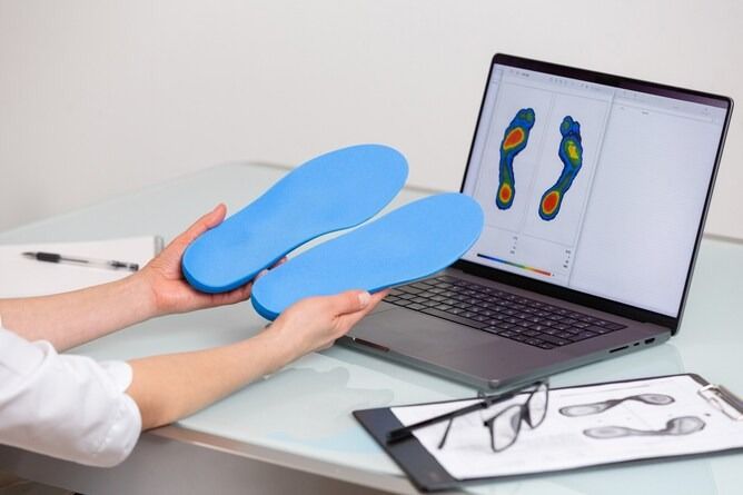 Una persona tiene in mano due solette ortopediche blu davanti a un computer portatile che visualizza una mappa della pressione plantare.