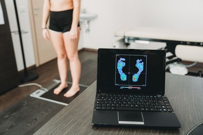 Una persona è in piedi su una pedana per la scansione podologica mentre i dati relativi alla pressione esercitata sul suo piede vengono visualizzati sullo schermo di un computer portatile in primo piano.