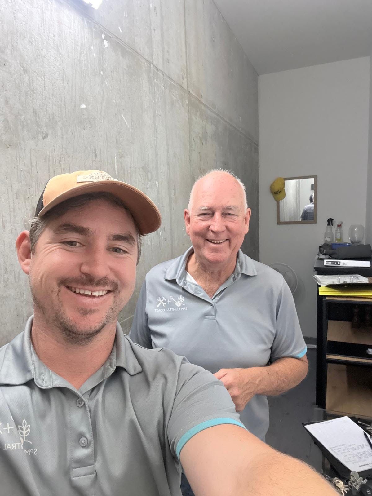 Two smiling people in a gray room, one taking a selfie near a desk and wall mirror.