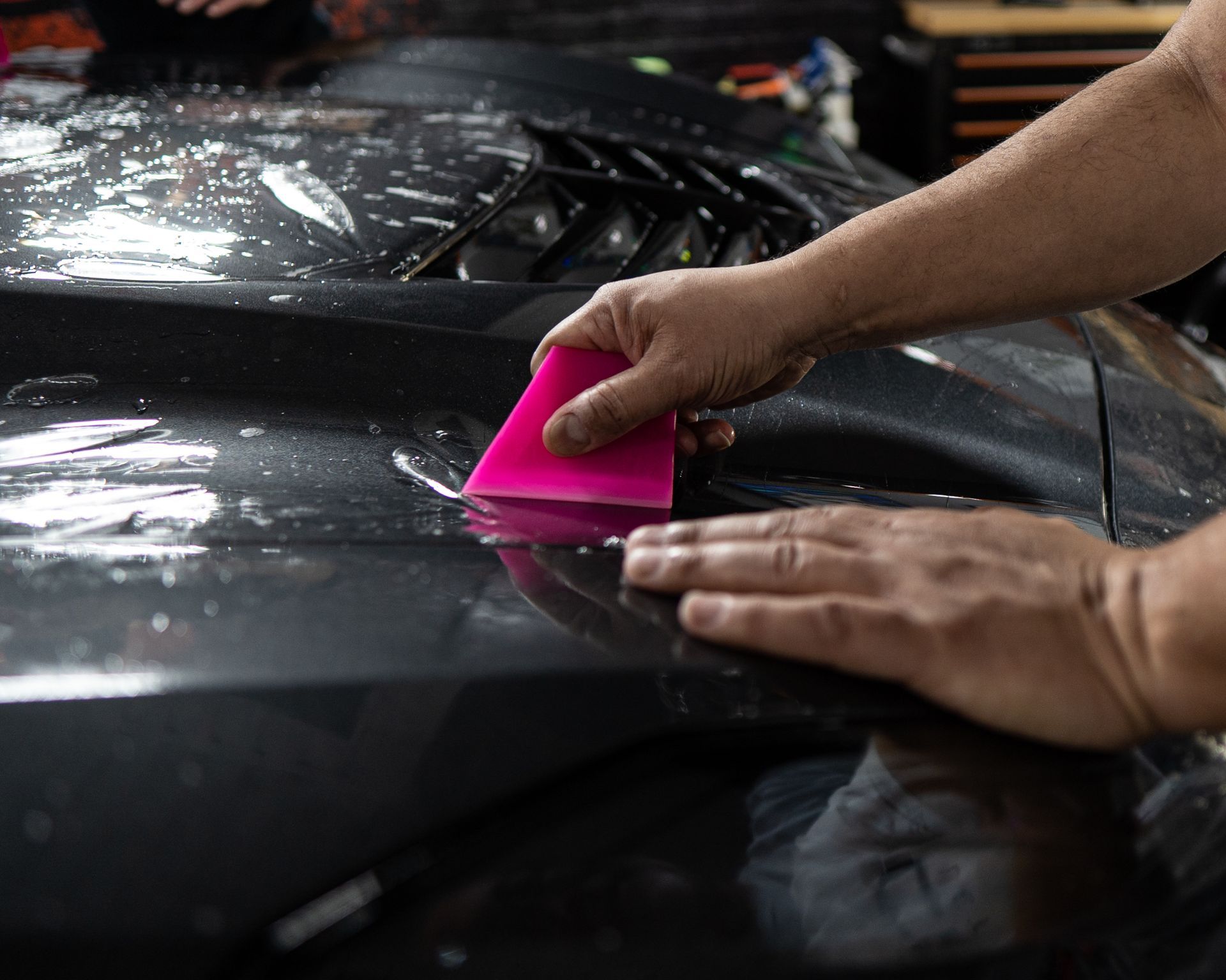 Hands applying protective film to a car hood using a pink squeegee.
