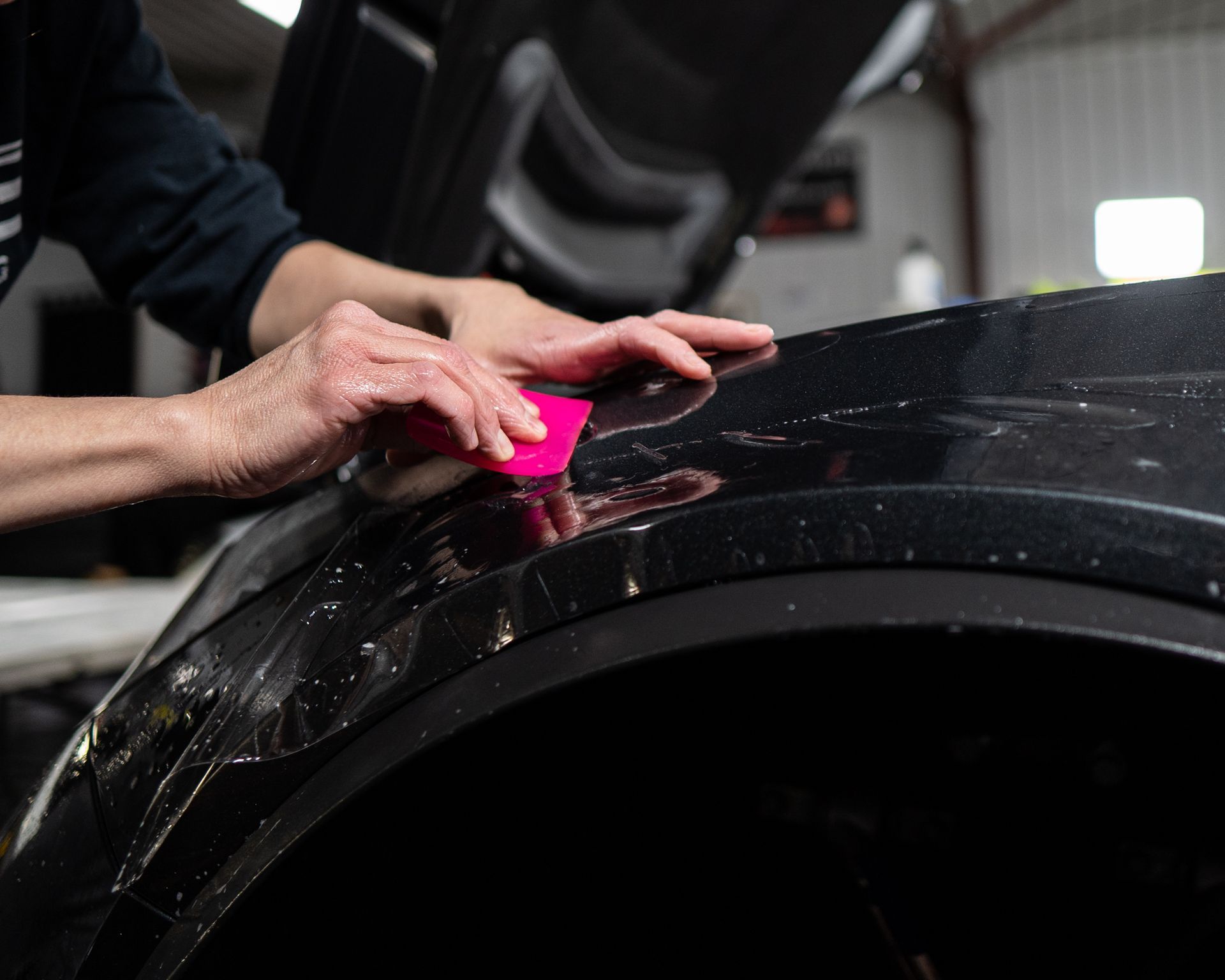 Person applying protective film to a black car fender with a pink squeegee in a workshop.