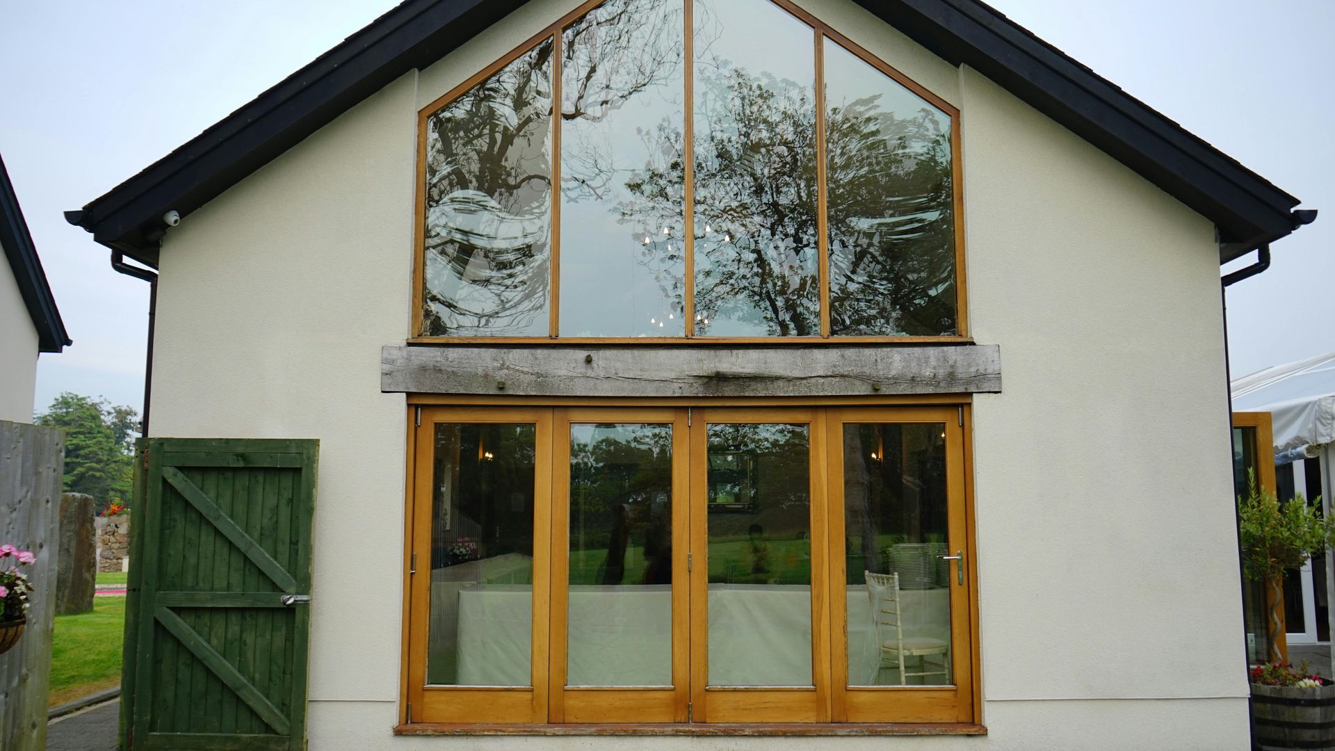A building with a large glass window, wooden doors, and green side door, reflecting trees and sky.