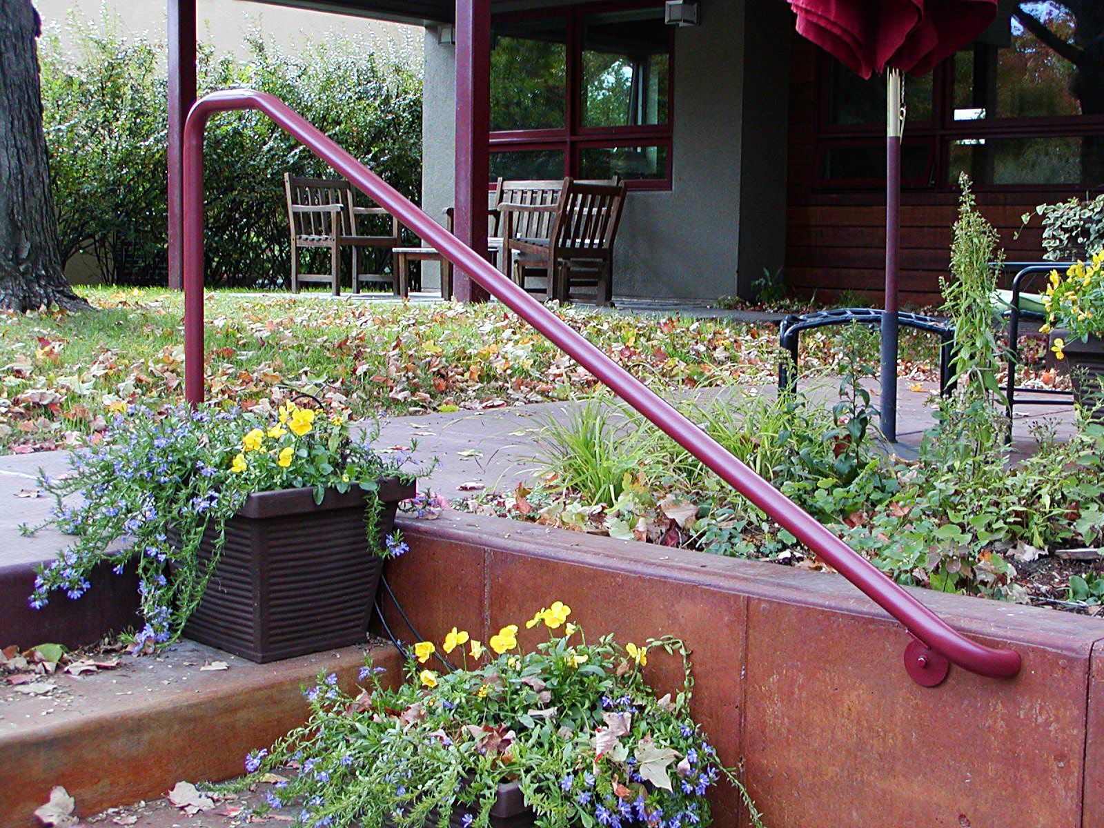 Red handrail on a low wall next to steps with potted flowers; a building in the background.