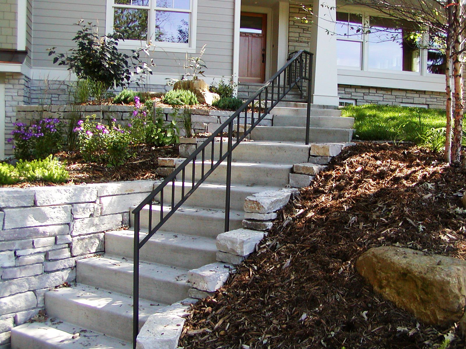 Concrete steps with a black handrail leading up to a house with landscaping and a stone retaining wall.
