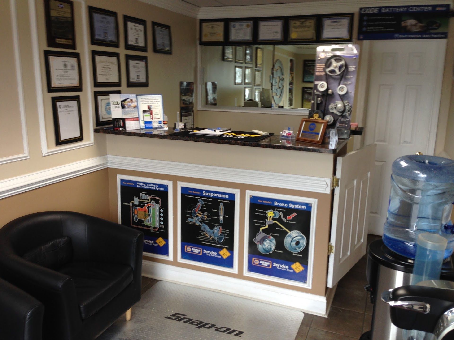 Office reception desk with framed certificates, awards, black chair, and water cooler under a wall mirror | Auto Clinic Care