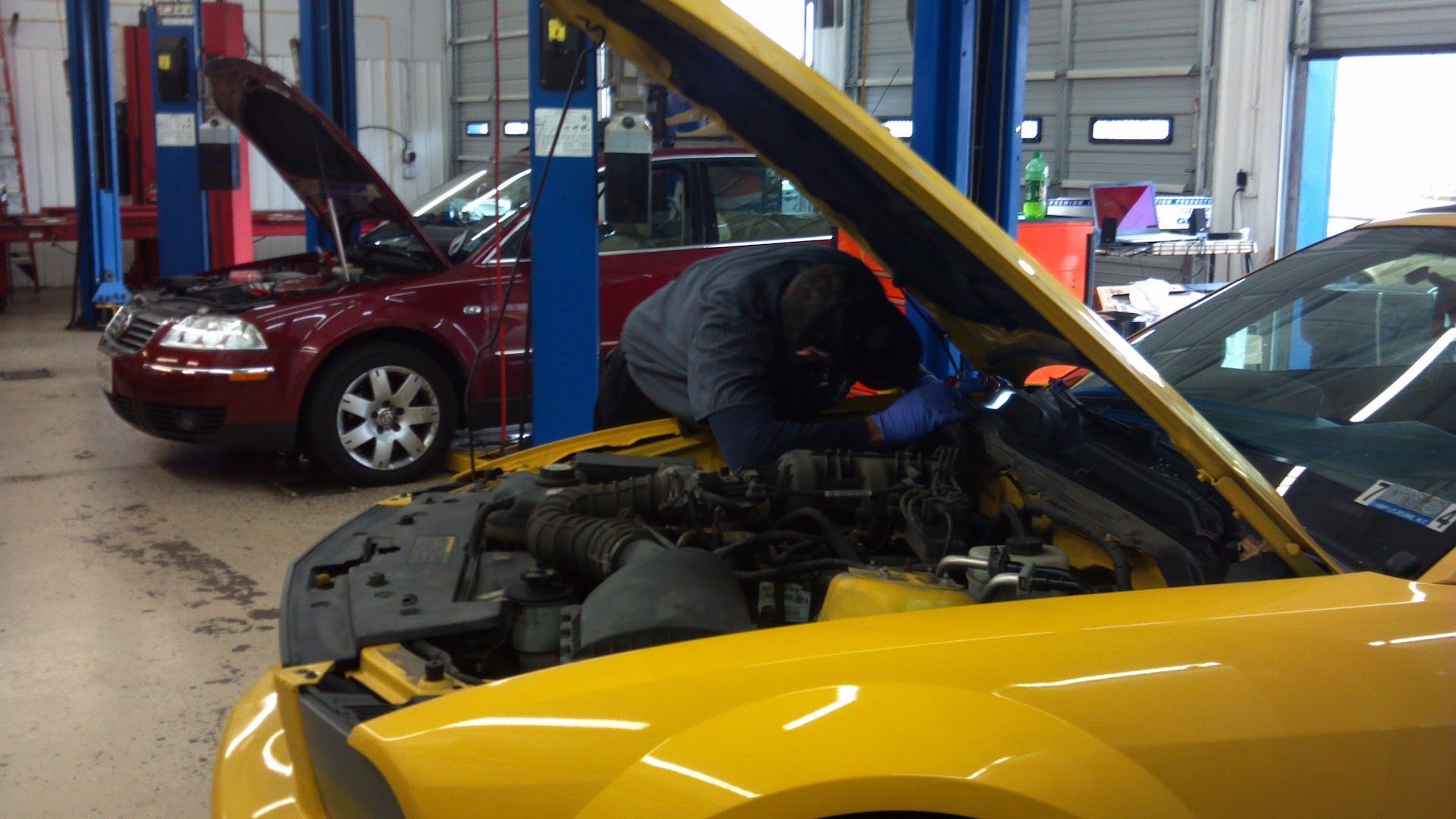 Mechanic working under the hood of a yellow car in an auto repair shop, with a red car in the background | Auto Clinic Care