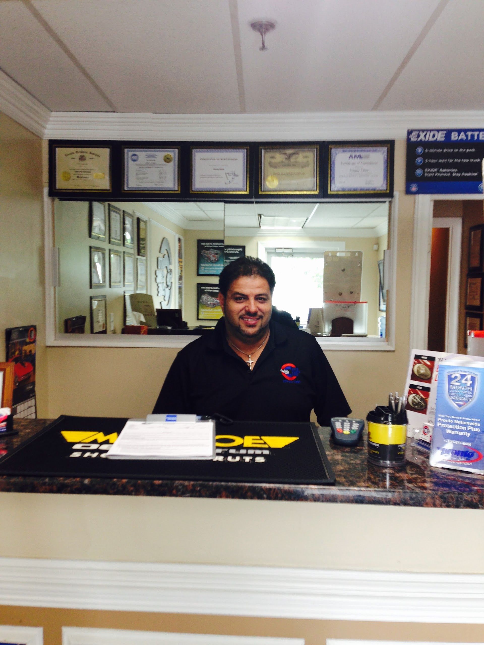 Hotel front desk clerk standing behind a counter in a lobby with framed notices and a mirror behind him | Auto Clinic Care