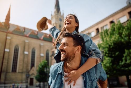 Man giving a woman a piggyback ride, both laughing outdoors near a church.
