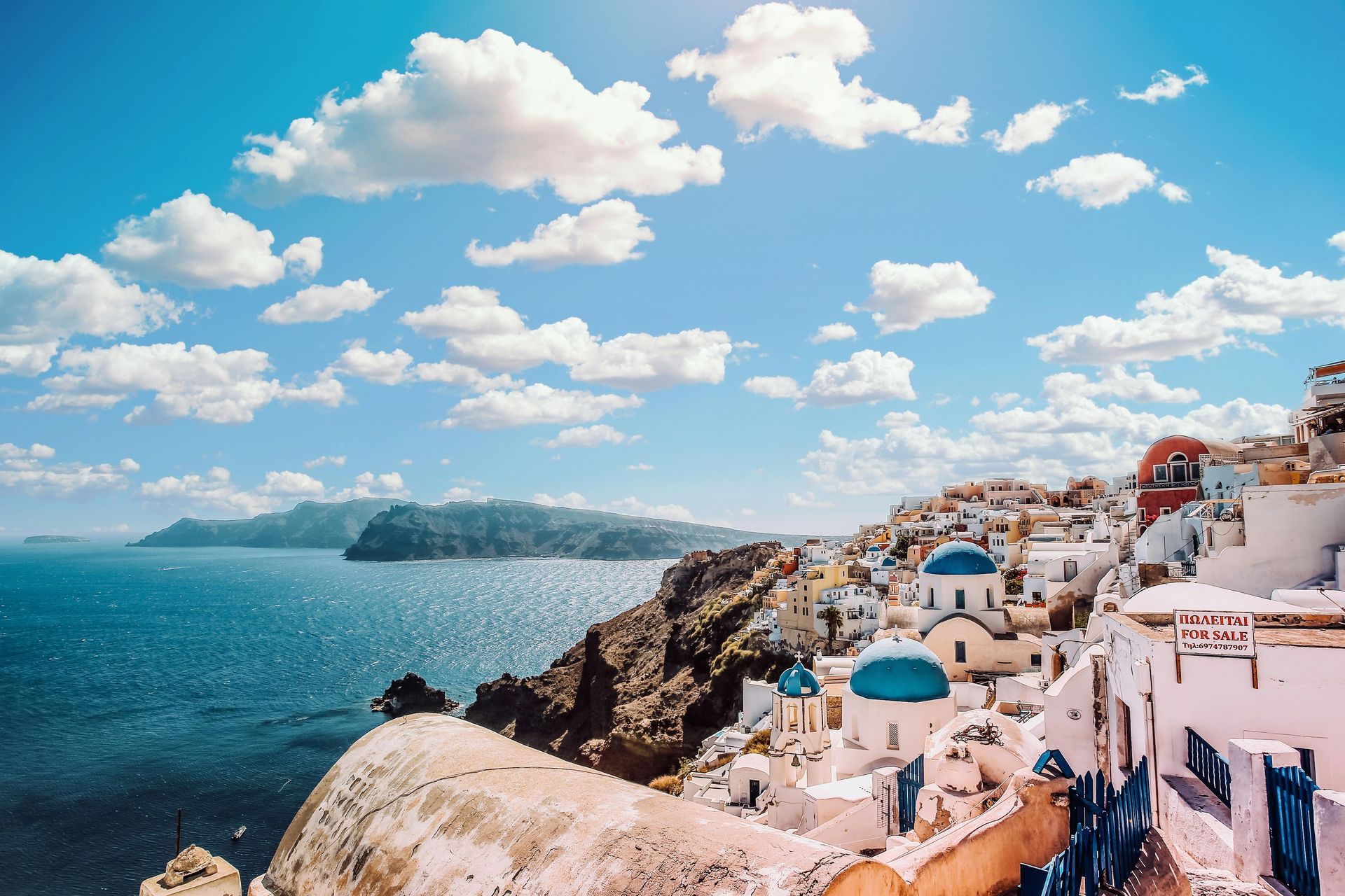 Coastal town in Greece with white buildings, blue domes, and sea under a bright blue sky with clouds.