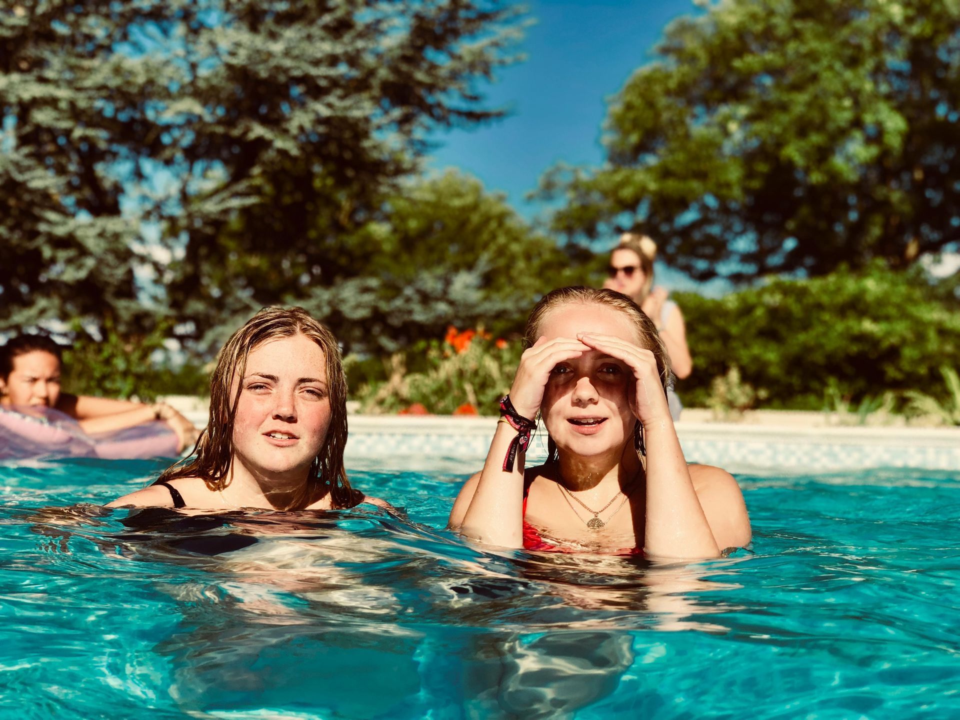 Two girls in a swimming pool on a sunny day. One shields her eyes, the other smiles. Trees in the background.
