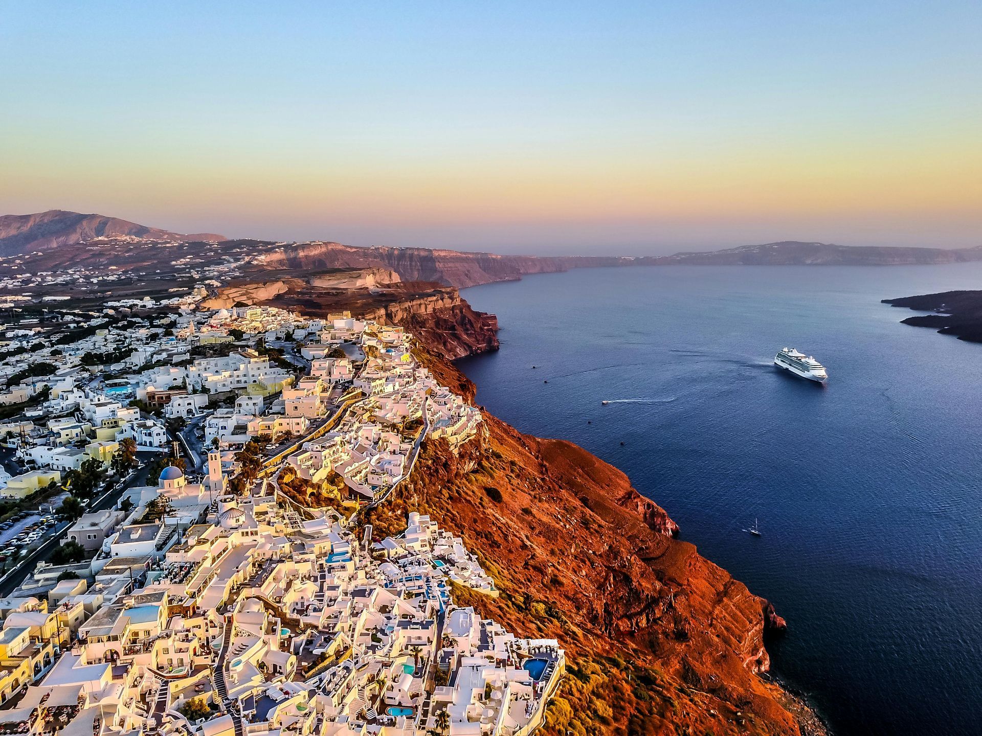 White buildings on a cliff overlooking the Aegean Sea, Santorini, Greece, with a cruise ship and sunset sky.