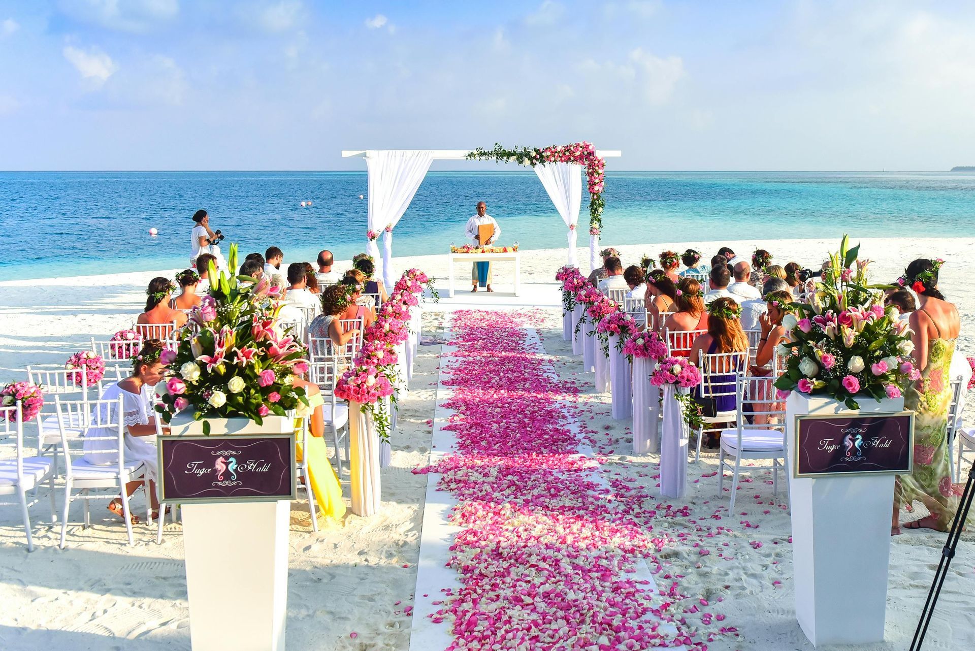 Beach wedding ceremony with pink floral aisle, white chairs, and ocean backdrop.