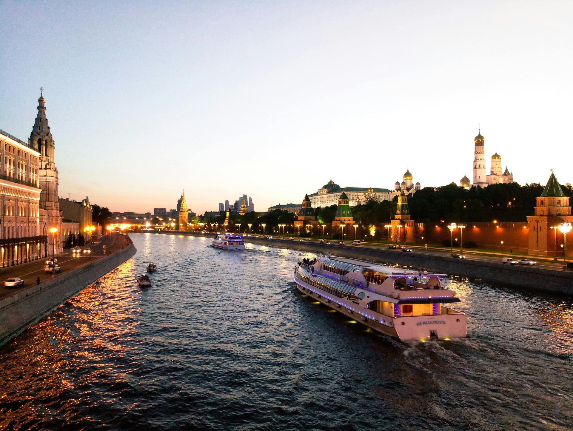 Boats on river, passing illuminated buildings including the Kremlin, Moscow, at dusk.