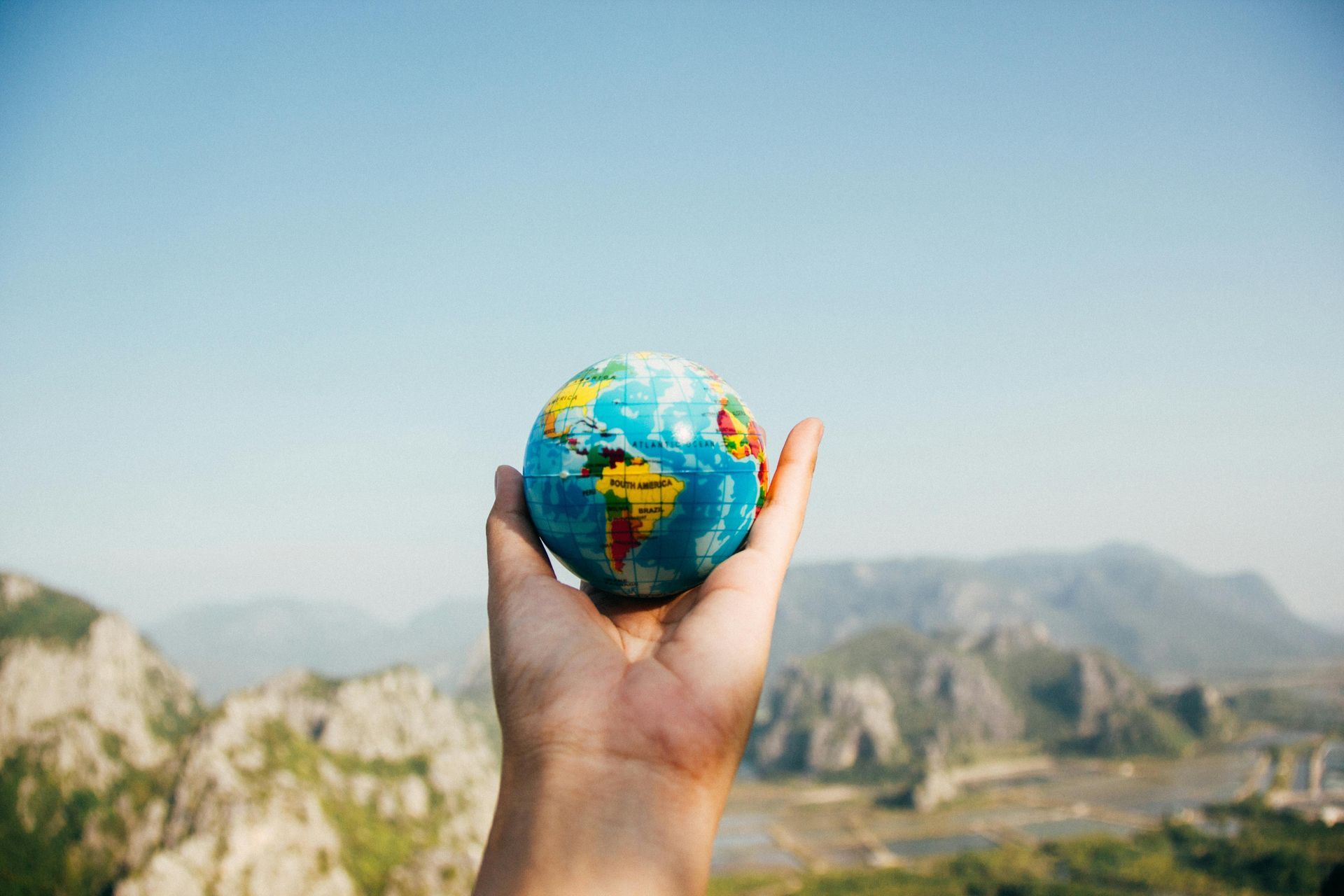 Hand holding a globe against a mountainous backdrop and clear sky.