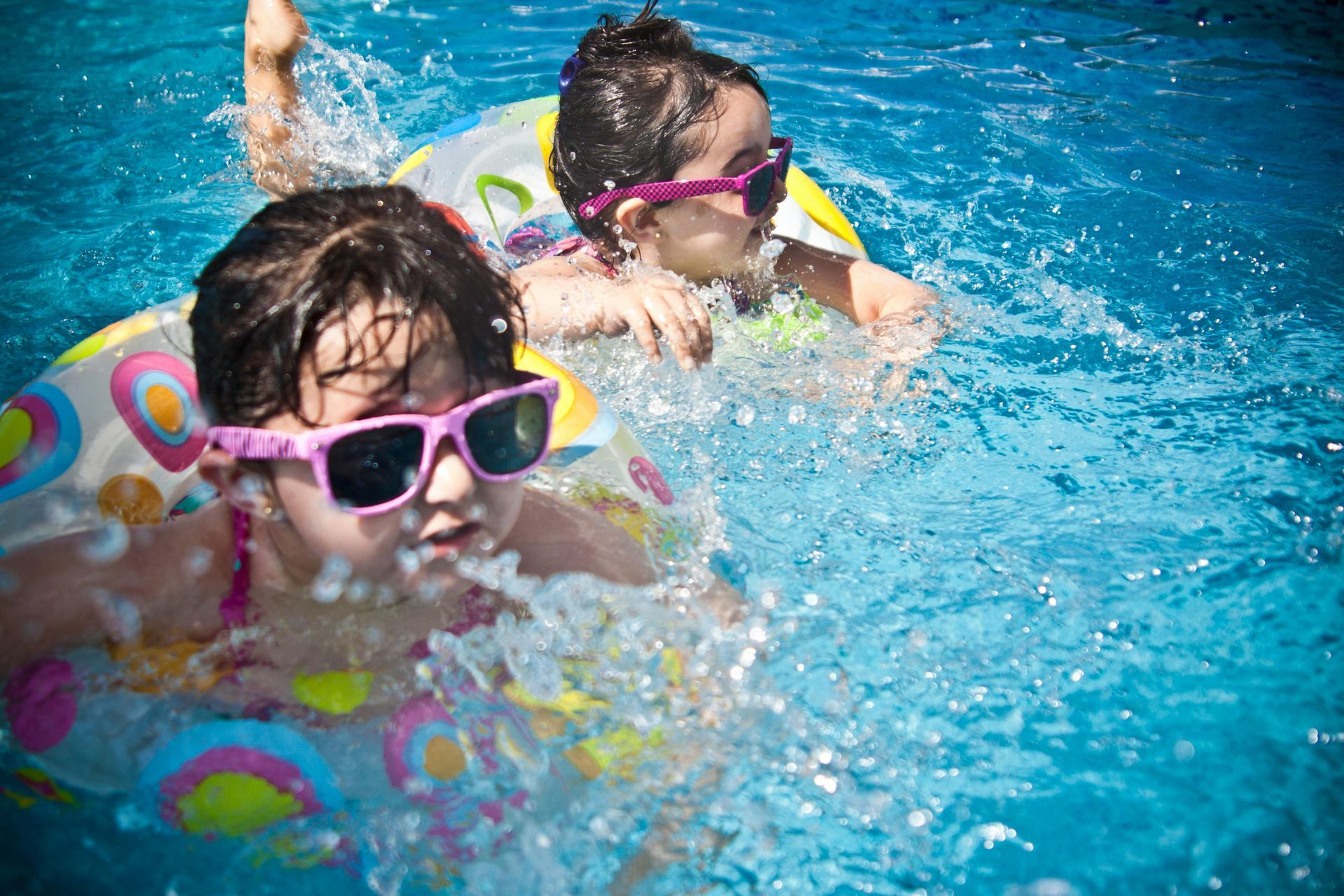 Two children in sunglasses swimming in a blue pool with inflatable rings.