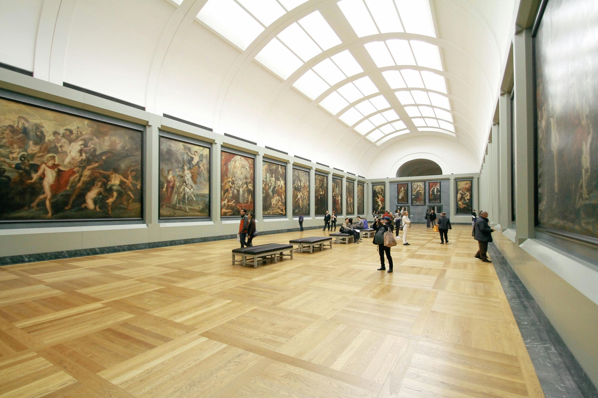 Museum interior with large paintings on walls, people viewing art, and a wood floor.