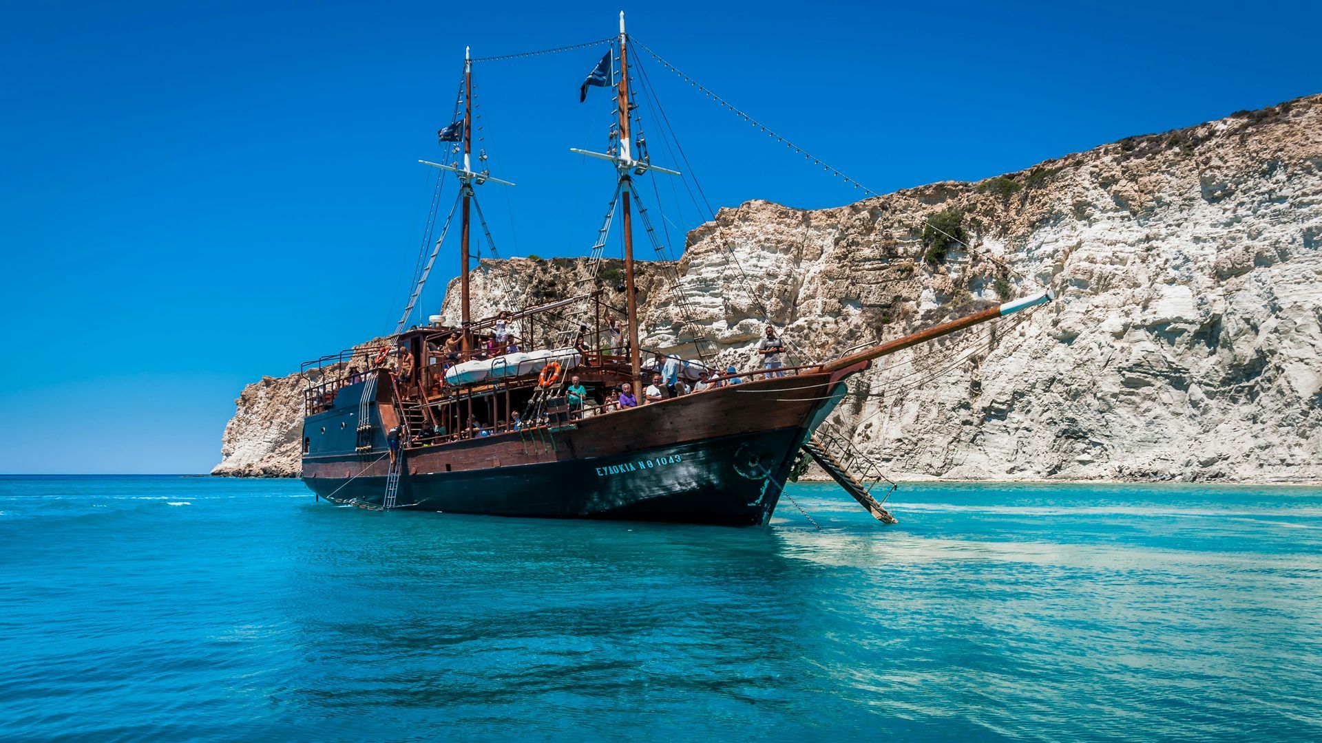 Pirate ship on turquoise water near a rocky cliff under a clear blue sky.