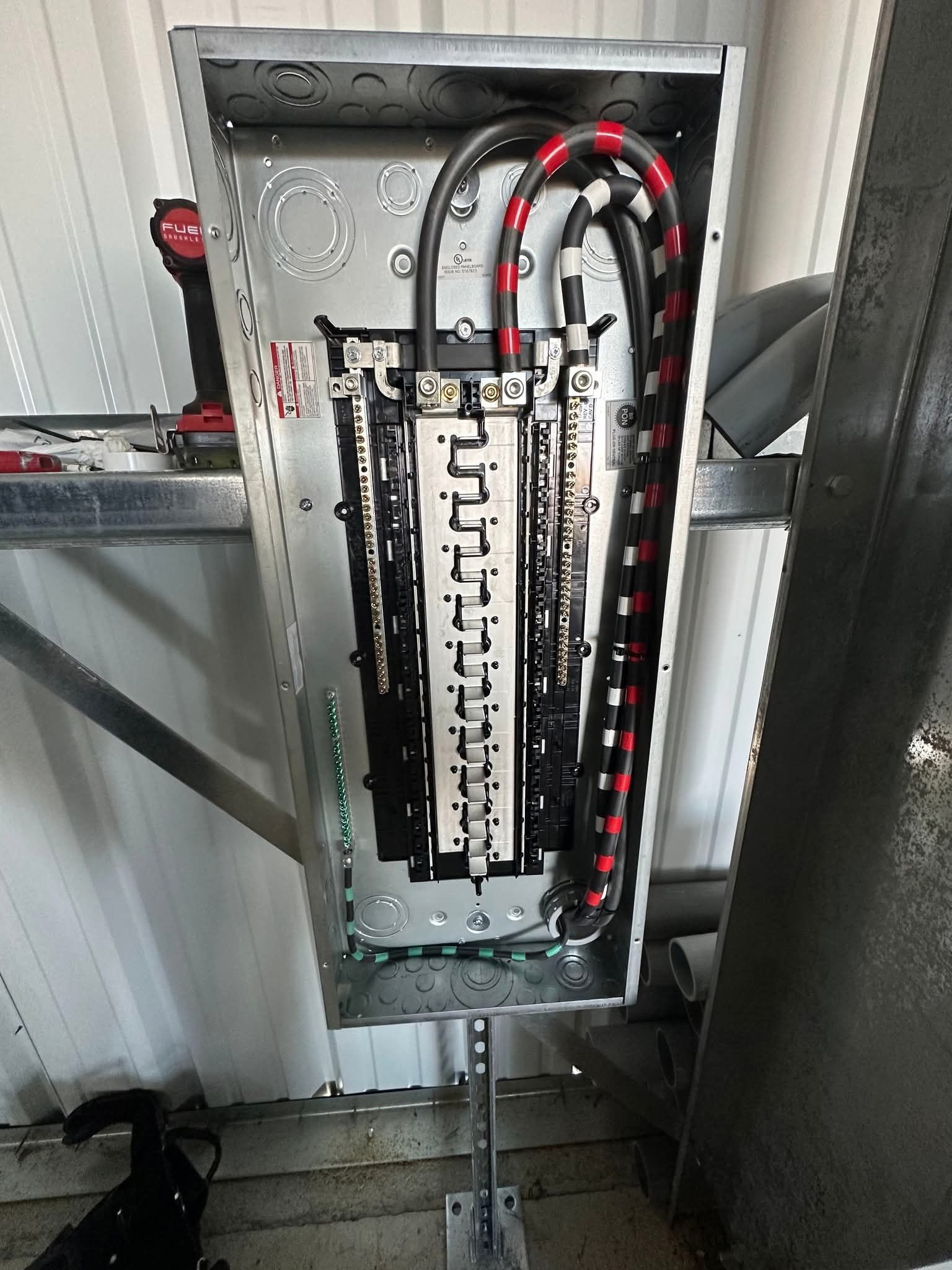 Electrical panel with black wires, installed on a metal bracket against a white wall.