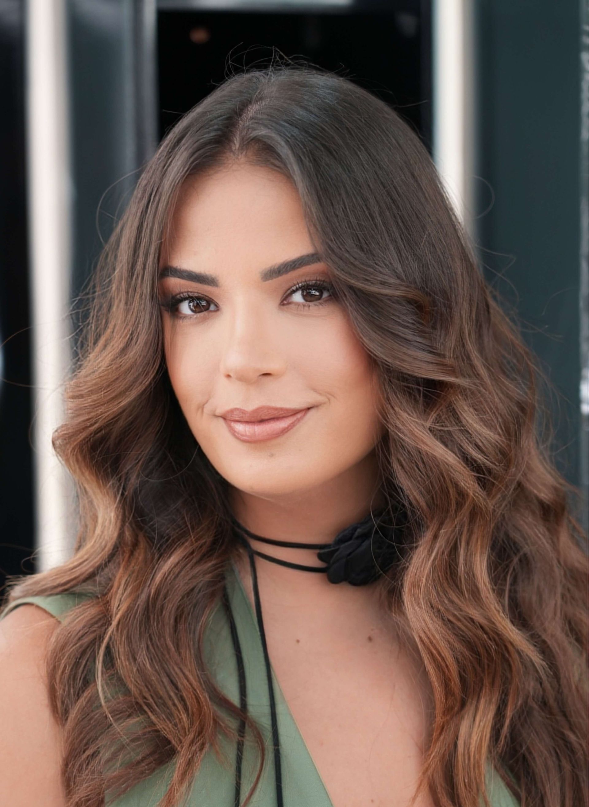 Woman with long, wavy brown hair smiles at the camera, wearing a black choker and green top.