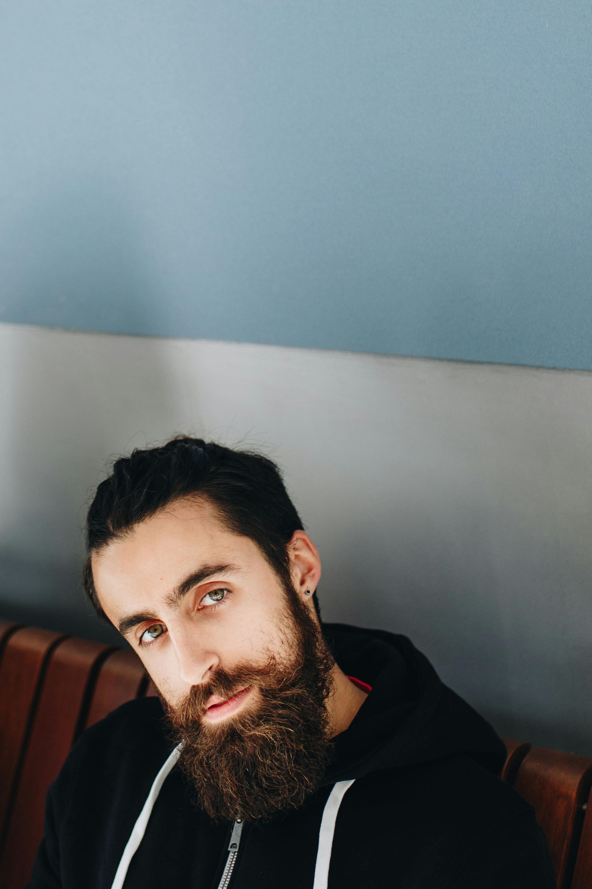 Man with a full beard, wearing a black hoodie, looking at the camera. He's sitting in front of a blue and gray wall.