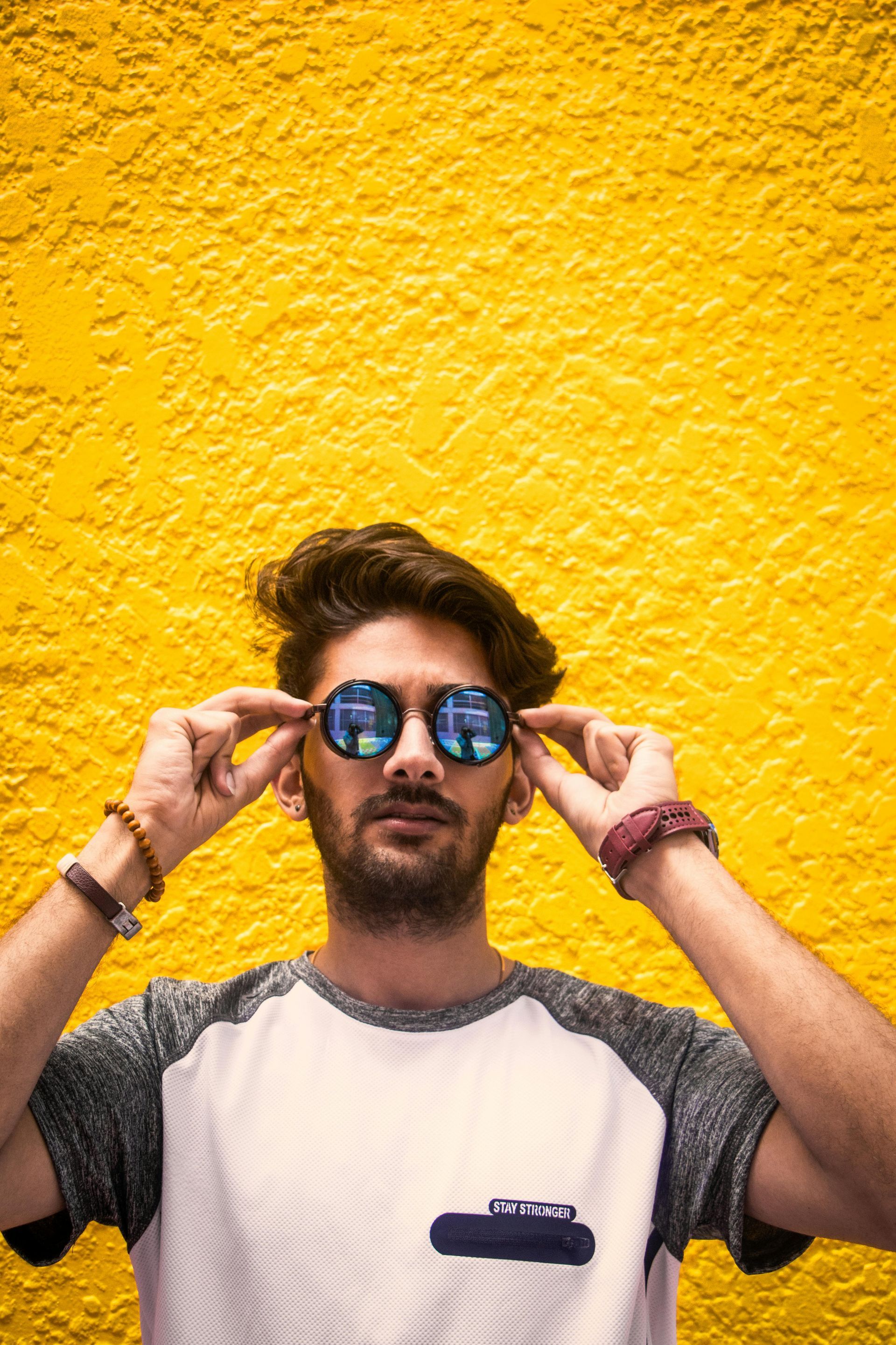 Man in sunglasses with a yellow textured wall background, adjusting glasses with both hands.