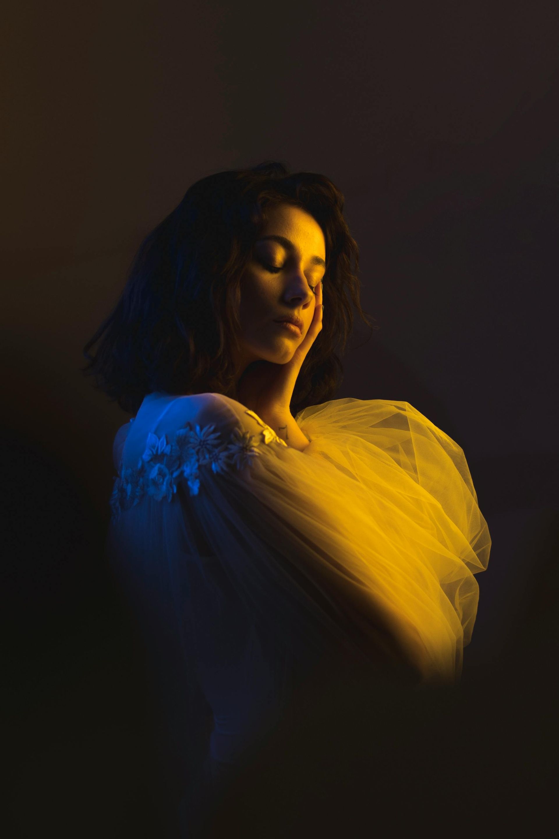 Woman in white dress, eyes closed, hand on cheek. Lit with blue and yellow light.