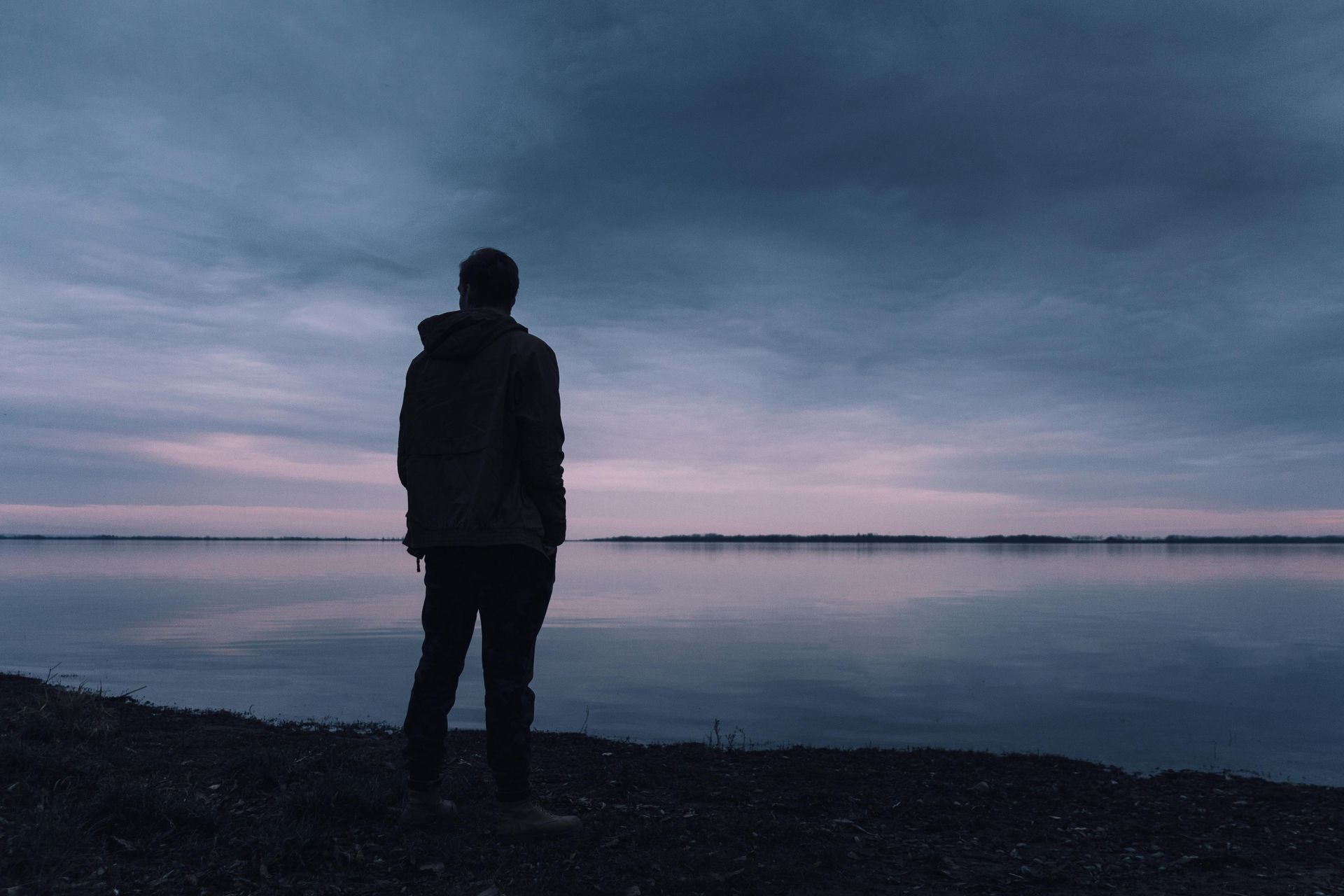 Person standing on a shore, facing still water under a cloudy sky at dusk.