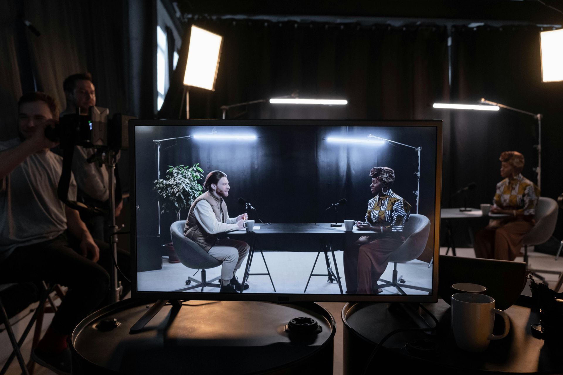 TV studio: A crew films a talk show with two guests at a table; view through a monitor.