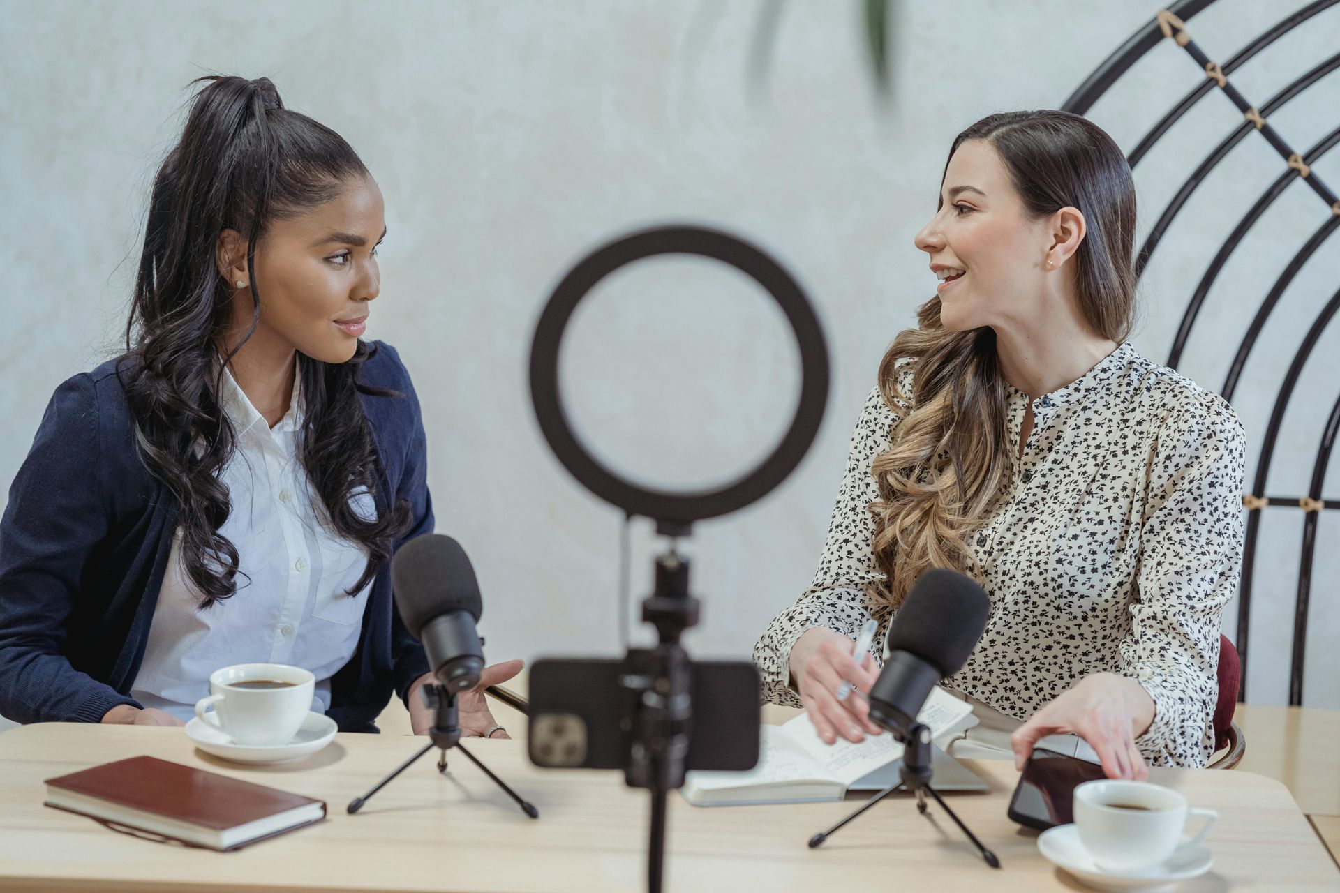 Two women podcasting at a table with microphones, ring light, coffee, and notebooks.