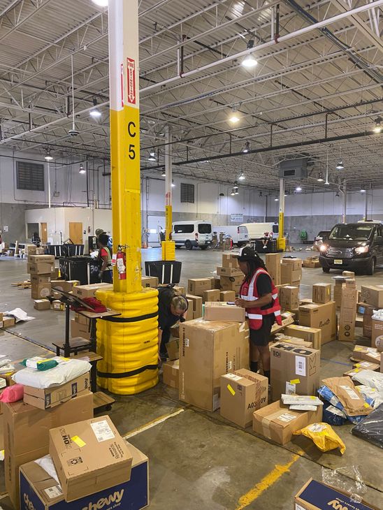 Warehouse with cardboard boxes, workers sorting packages, and a yellow support pillar under bright lights.