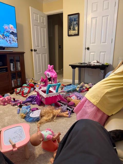 A living room floor covered with scattered toys, dolls, and a toy cart, showing how hard it is to find one item in a cluttered space