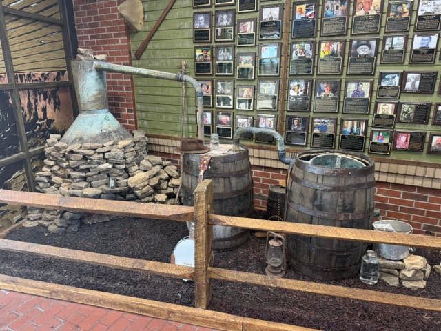 A copper moonshine still with a condenser pipe standing in front of a wall of framed plaques at the Dawsonville Moonshine Distillery