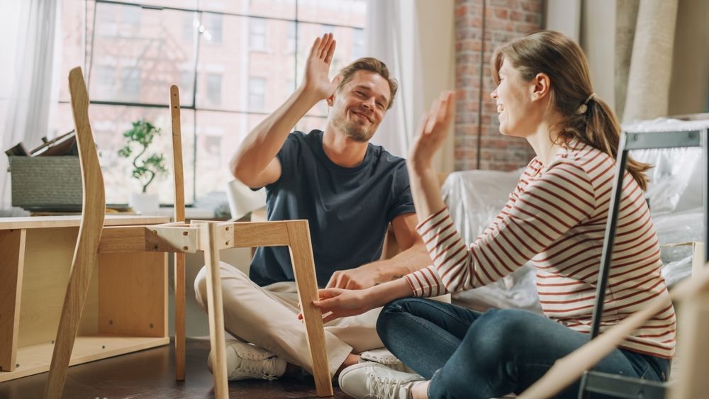 Couple assembling furniture, high-fiving in a new apartment, joyful expressions, sunny window, light wood chairs.
