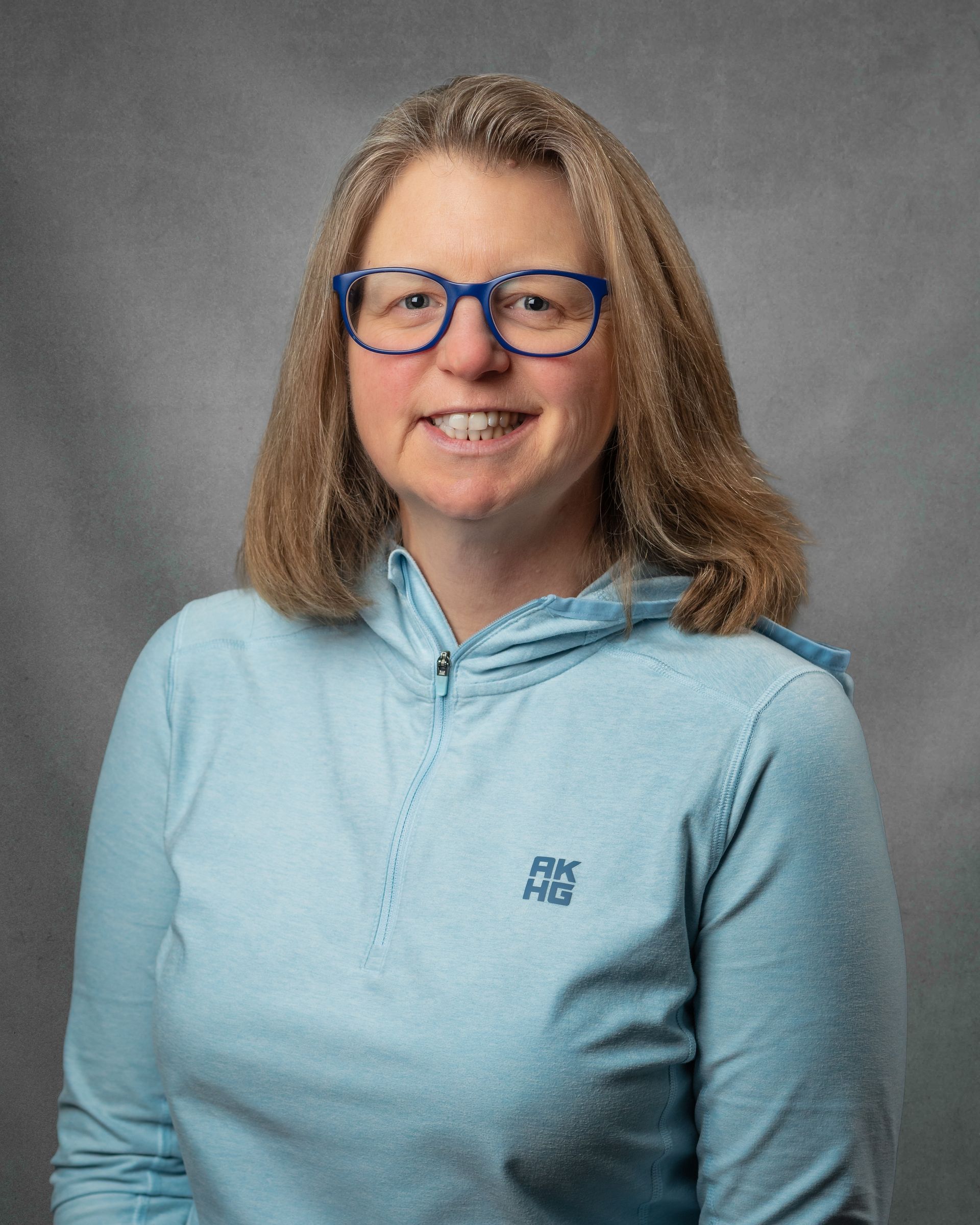 A woman wearing glasses and a blue shirt is smiling in a hallway.