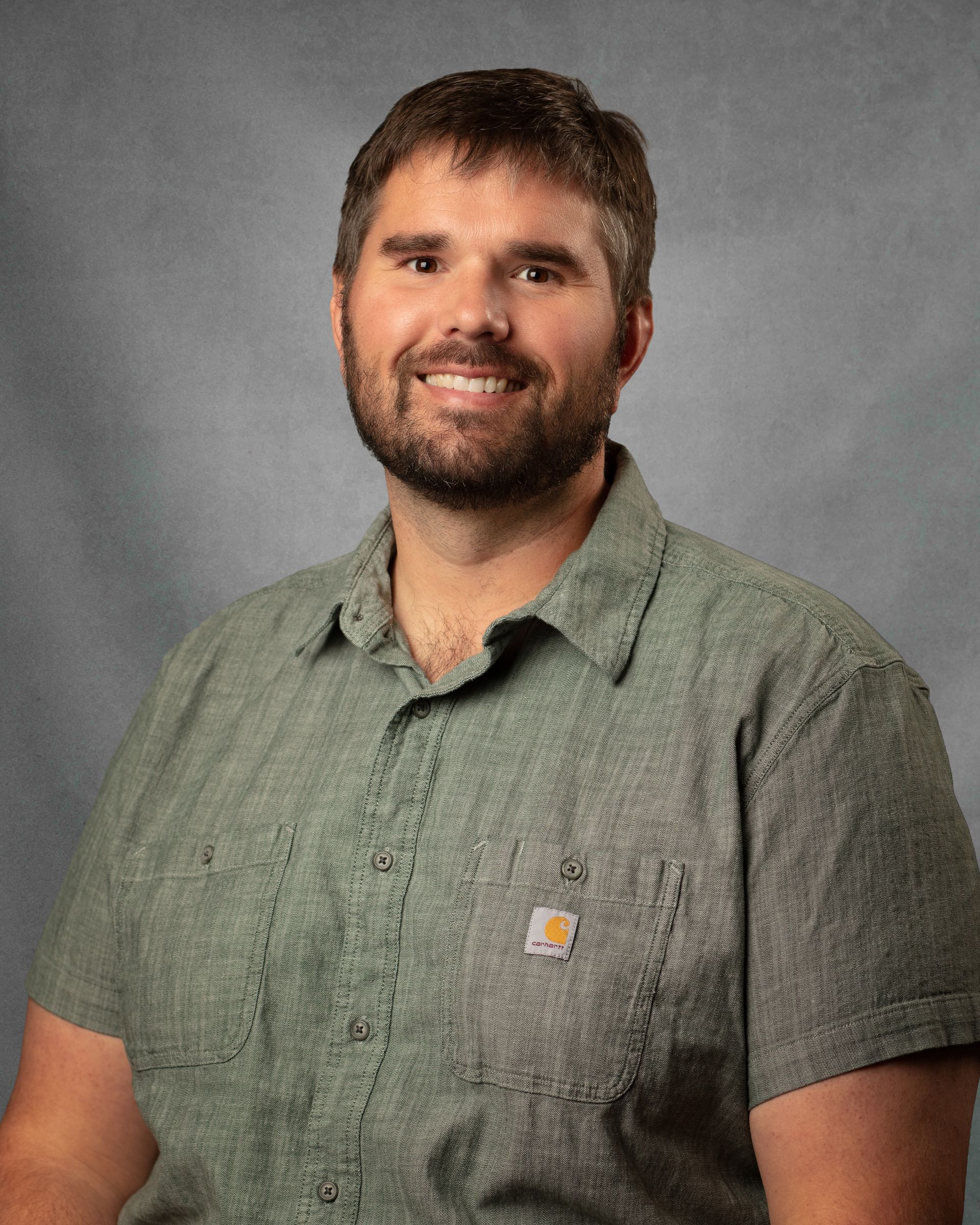 A man with a beard wearing a green shirt is smiling in front of a brick wall.