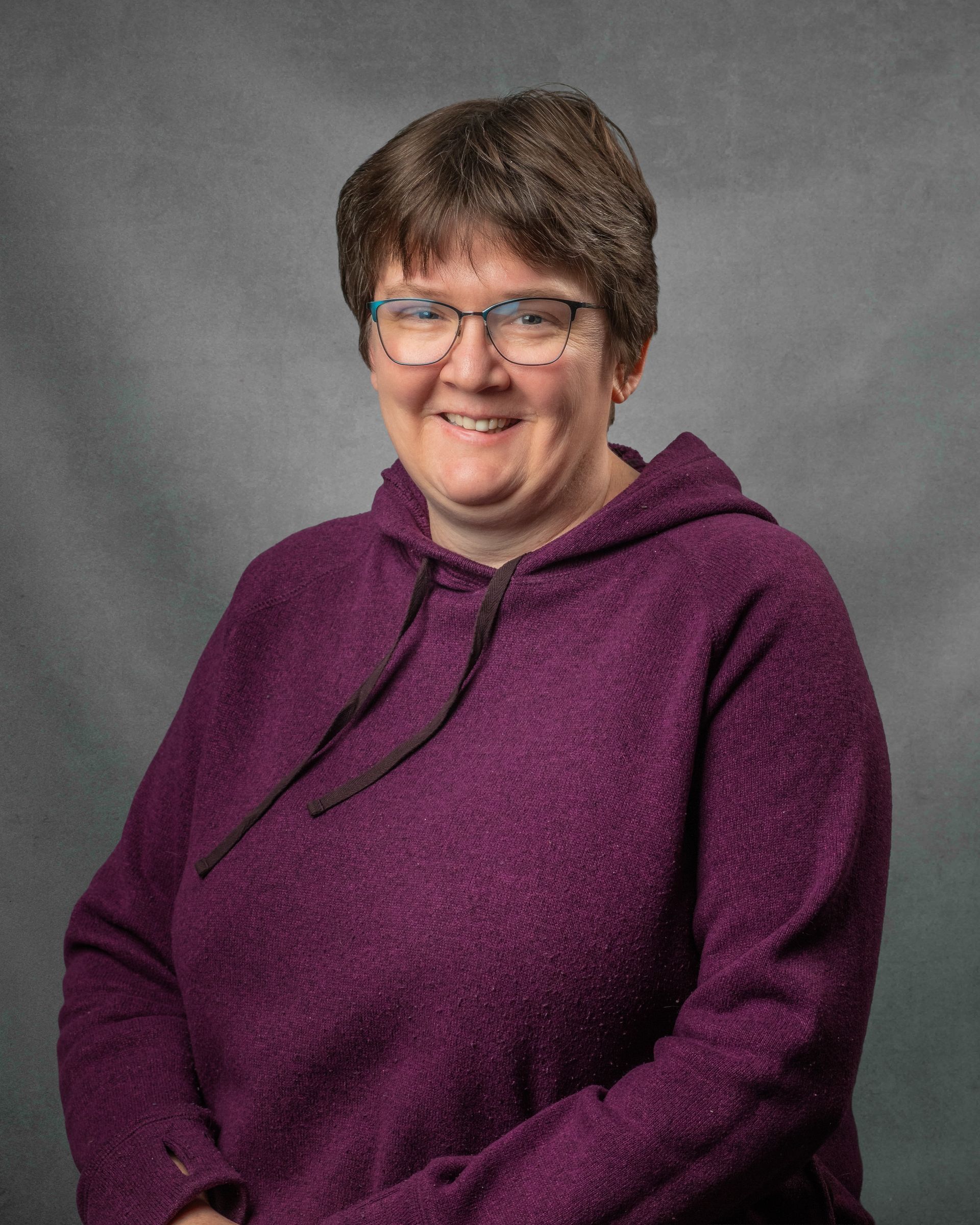 A woman wearing glasses and a purple sweat shirt is smiling for the camera.