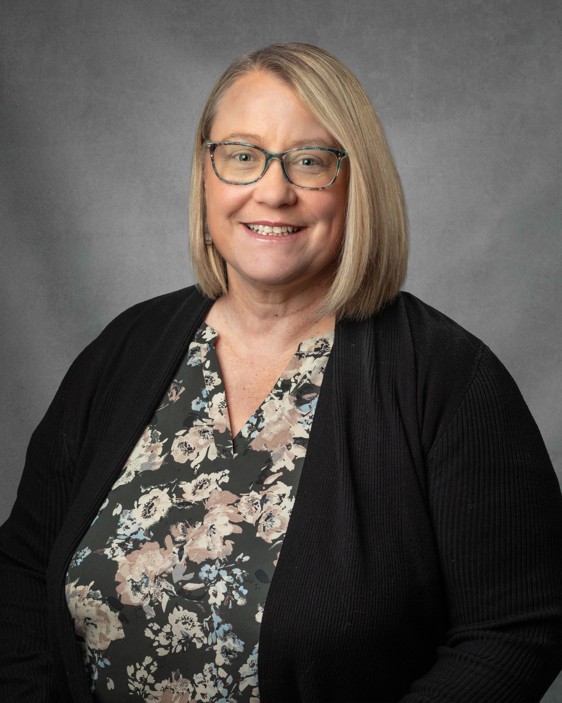 A woman wearing glasses and a floral top is smiling for the camera.