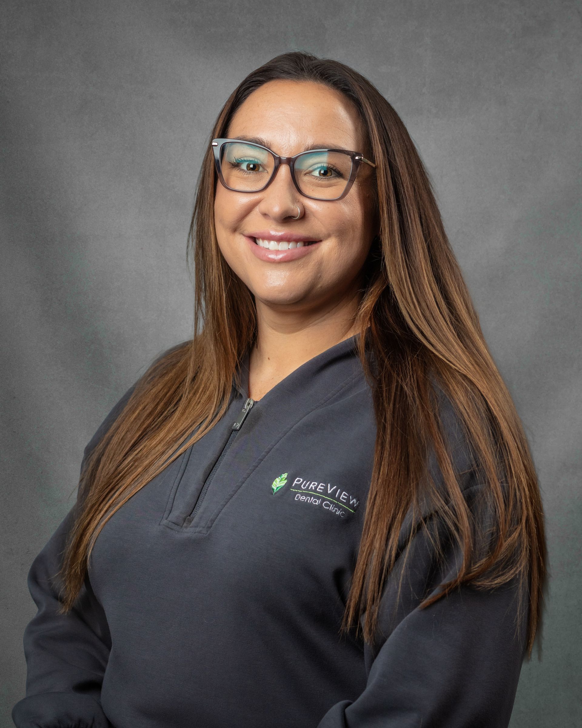 A woman wearing glasses and a gray zip up is smiling in a dental office